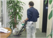 清掃会社の男性職員が長袖の青いジャンパーに白い長ズボンを身にまとい、ビル内のオフィスの緑の床を床洗浄機で清掃している。オフィスの壁は白く、テーブルは茶色い。床には、観葉植物や茶色いテーブルが置かれている。