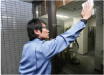 清掃会社の男性職員が長袖の青い作業着を身にまとい、オフィスの窓を布で拭いている。職員は黒髪で黒縁眼鏡を掛けている。オフィスの壁面は灰色のタイルである。
